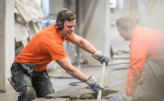 Två män i orangea tröjor och med hörselskydd arbetar vid en betongstation med betong. Den ena håller i en pinne och de båda ser glada ut. 