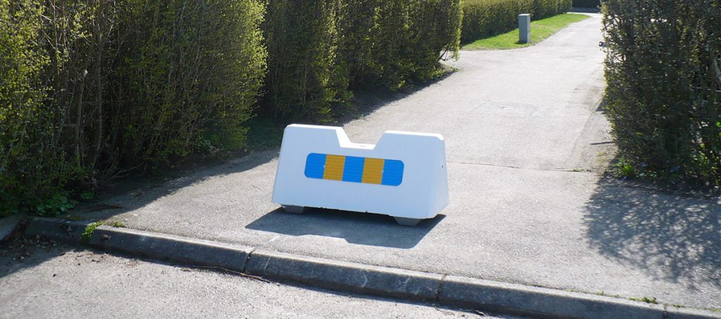 En vit trafikplint i betong med blå och gul reflex som står på en trottoar med gröna buskar runt omkring