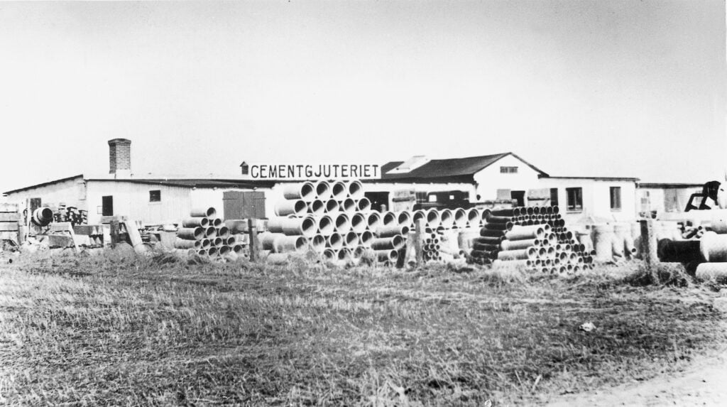 En äldre bild i svartvitt på cementgjuteriet med en text på taket där det står cementgjuteriet med stora bokstäver och där det ligger betongrör travade på marken i förgrunden.